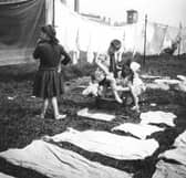 Children help each other clean up as they hang out washing on Glasgow Green. The square tower of the Greenhead Public Baths and steamie can be seen in the distance above the washing lines at Greenhead Public baths.